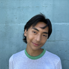 person with short dark hair, head slightly tilted, smiling at the camera. They are wearing a white tshirt and are standing in front of a light blue wall